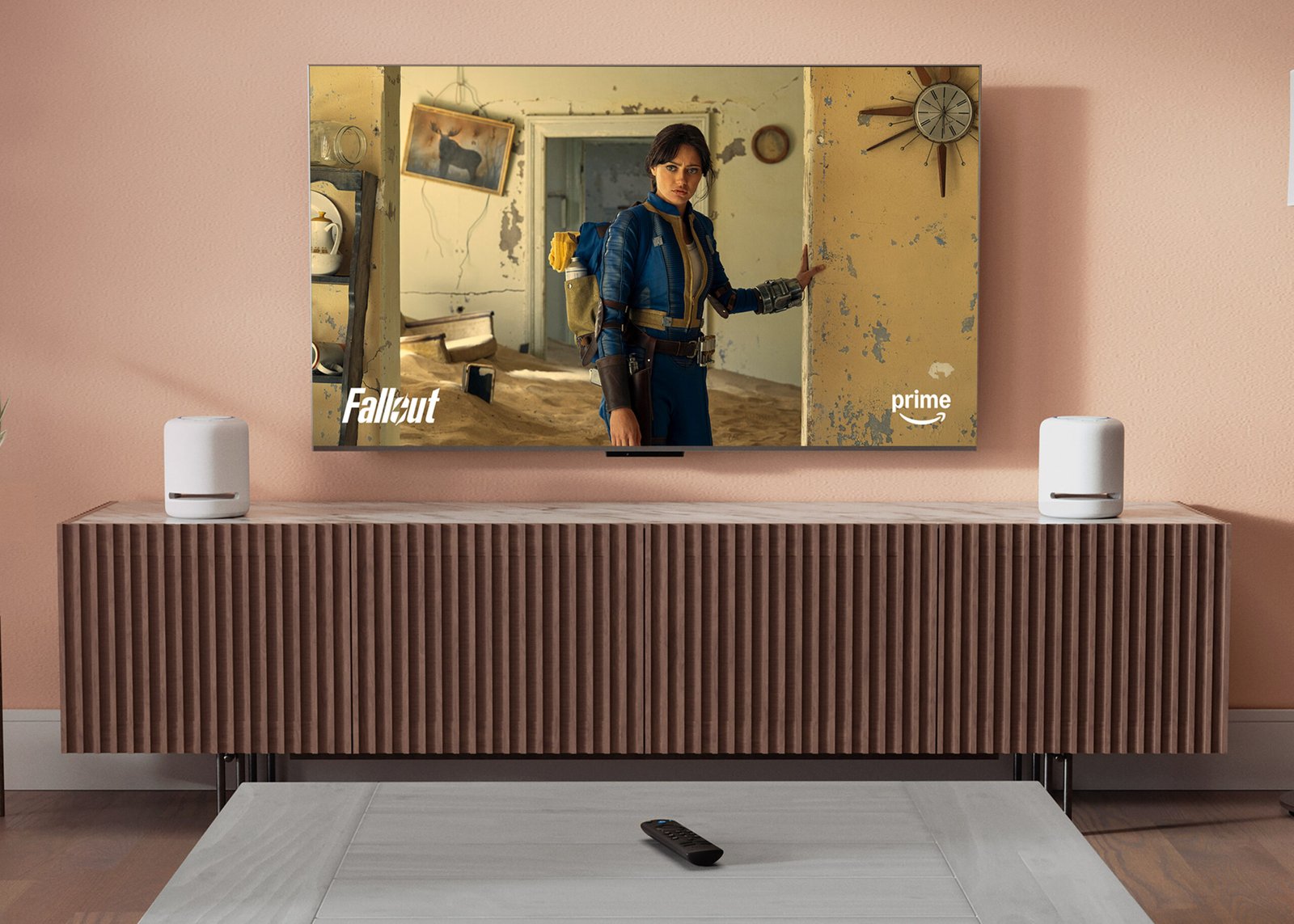 HDTV mounted on a wall in a modern living room showing Prime Video content, next to 2 Echo Studio 7-inch wide and 8-inch tall cylindrical mesh speakers with flat bottoms and tops, and 4 raised buttons on the top.